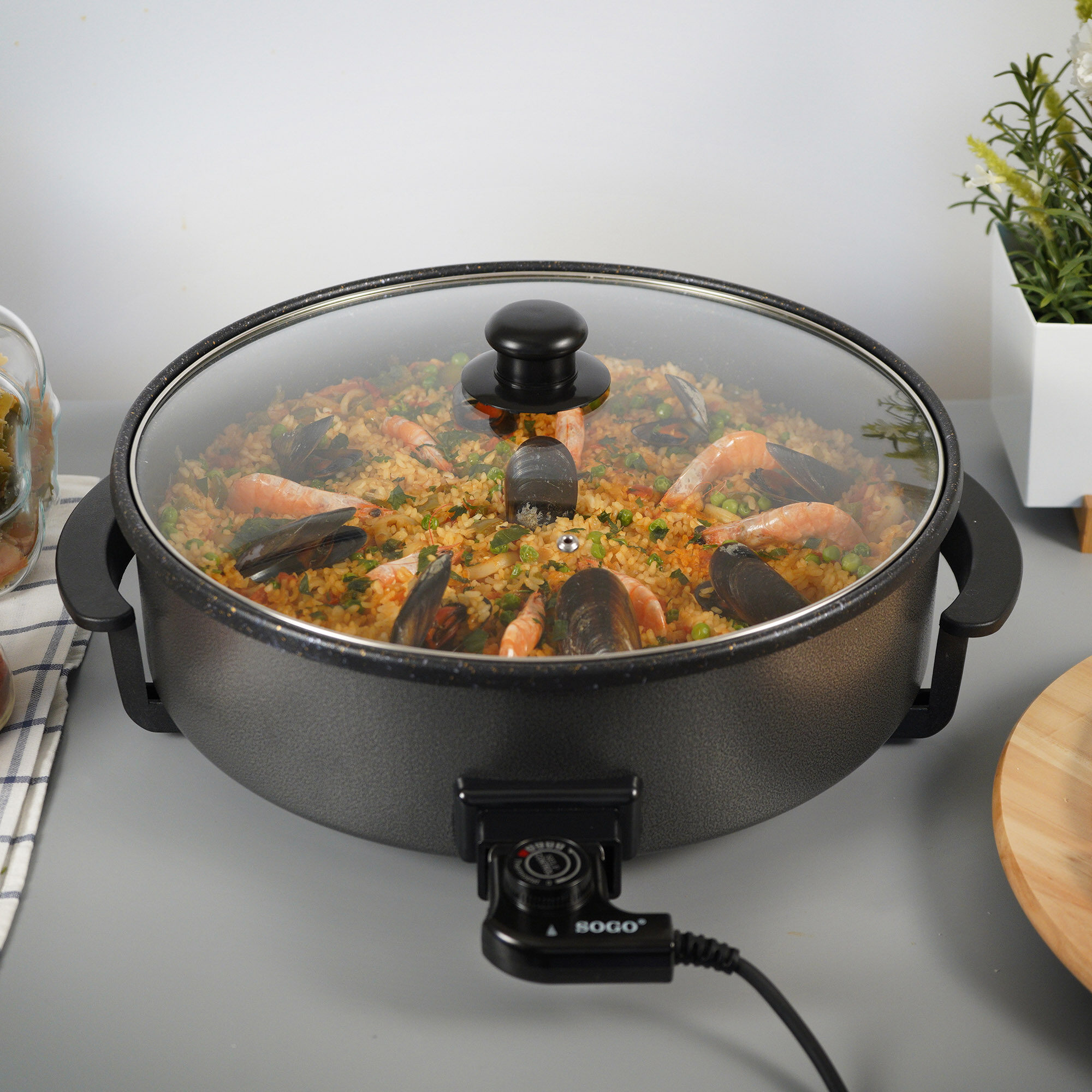Padella elettrica con paella, gamberi e cozze sotto un coperchio di vetro.