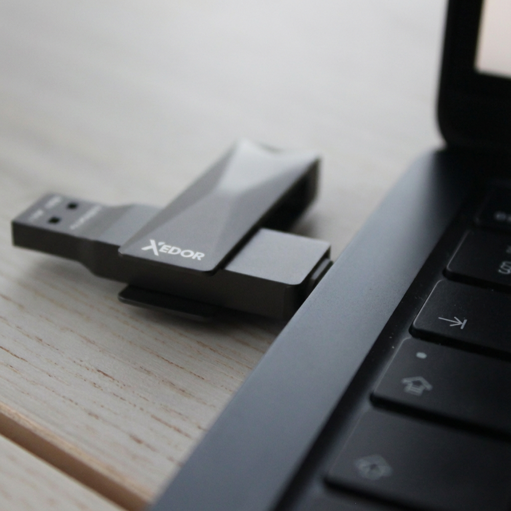 A gray XEDOR USB drive plugged into a laptop keyboard on a wooden surface.