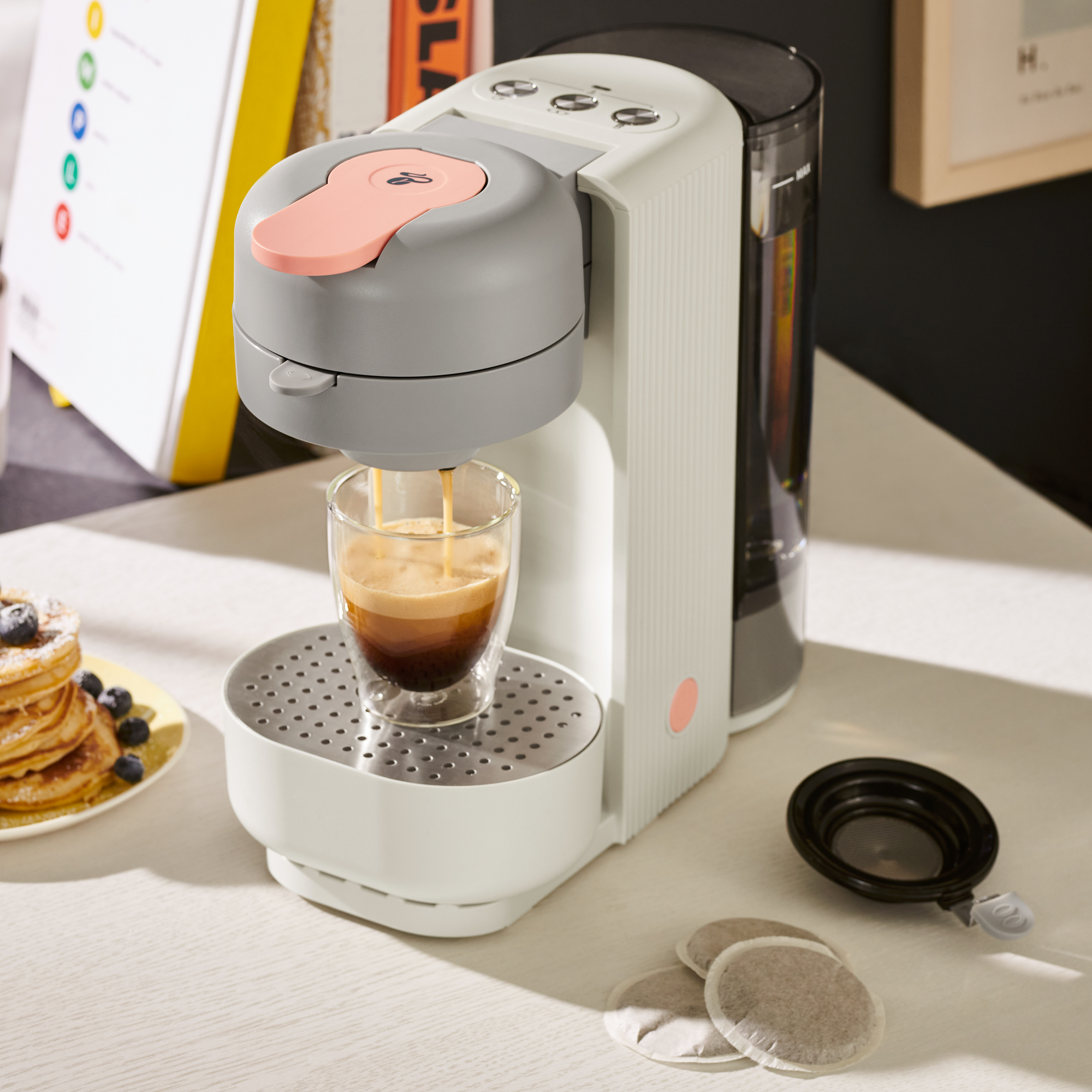 Eine weiße Kaffeemaschine brüht Kaffee in ein Glas, daneben stehen Pfannkuchen.