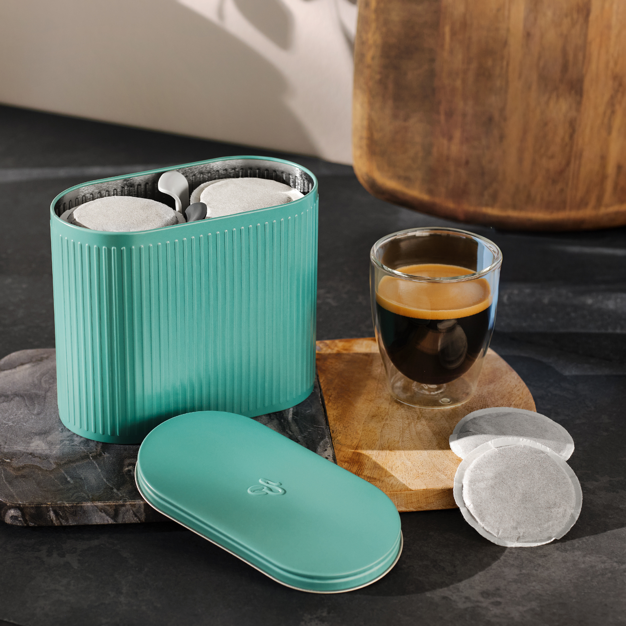 Türkiser Behälter mit Kaffee-Pads, daneben ein Glas Espresso und weitere Pads.