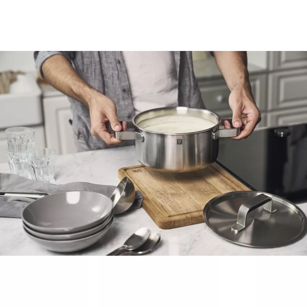 Hombre sujetando olla de sopa sobre tabla de cortar. Cuencos y cucharas cerca.