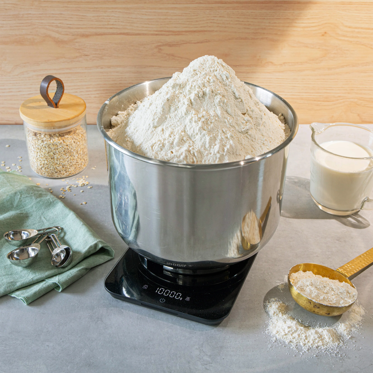 Ein Edelstahltopf voller Mehl steht auf einer Waage neben Haferflocken und Milch.