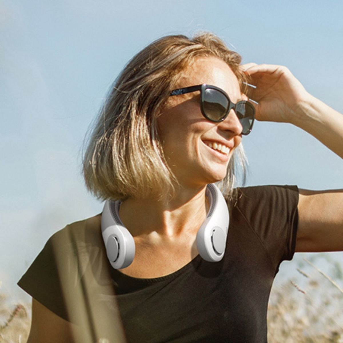 Frau mit Nackenventilator, Sonnenbrille, im Freien bei blauem Himmel.