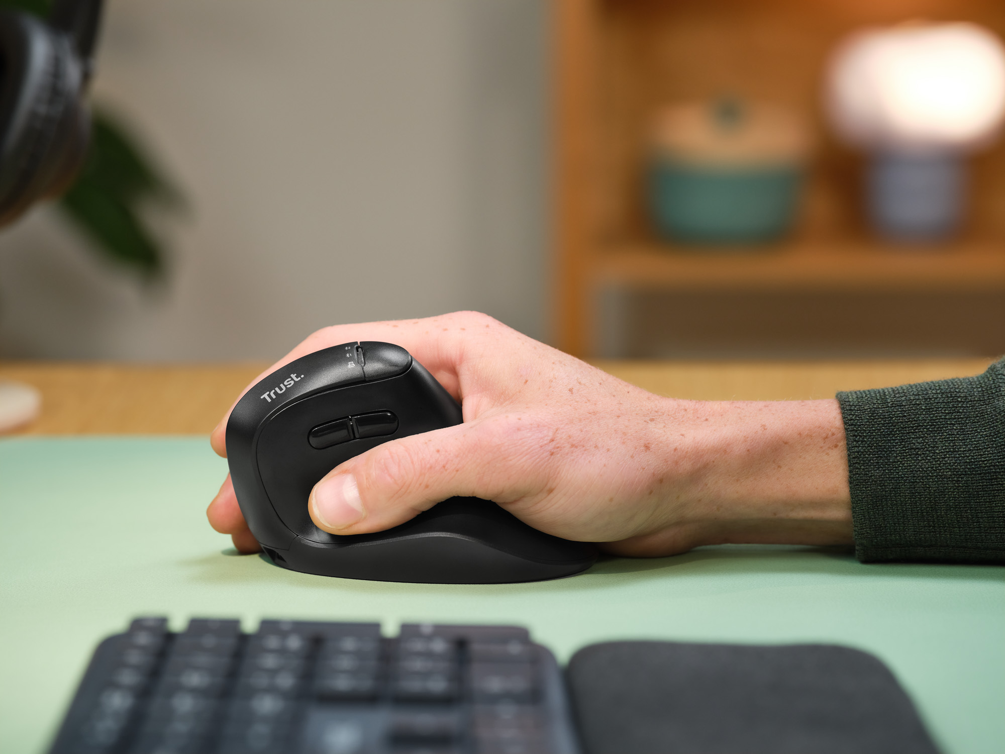 Une main manipule une souris noire sur un bureau vert clair. Un clavier est au premier plan.