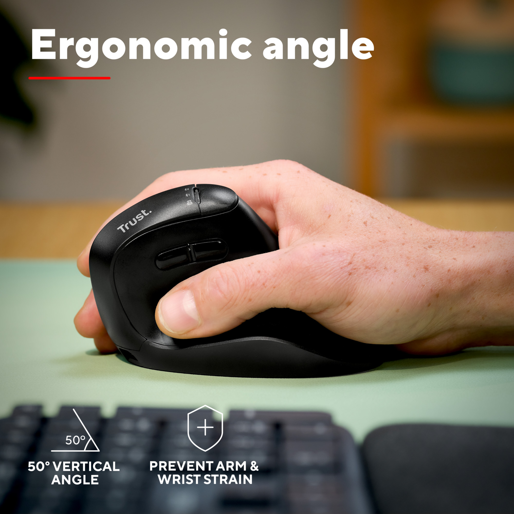 Main tenant une souris ergonomique noire avec l'étiquette 'Trust.', près d'un clavier.
