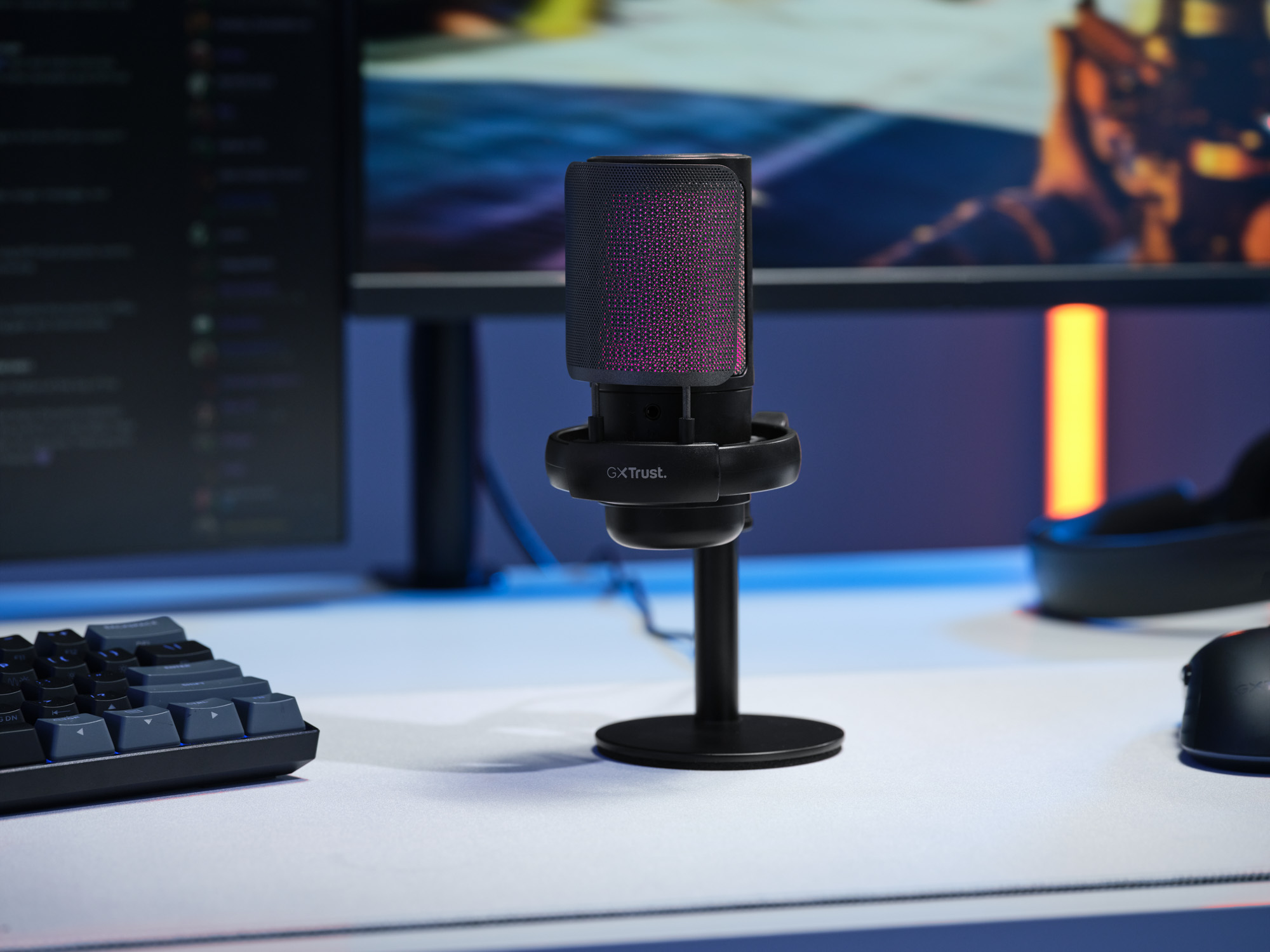 Un microphone Trust noir avec éclairage violet est posé sur un bureau avec clavier et souris.