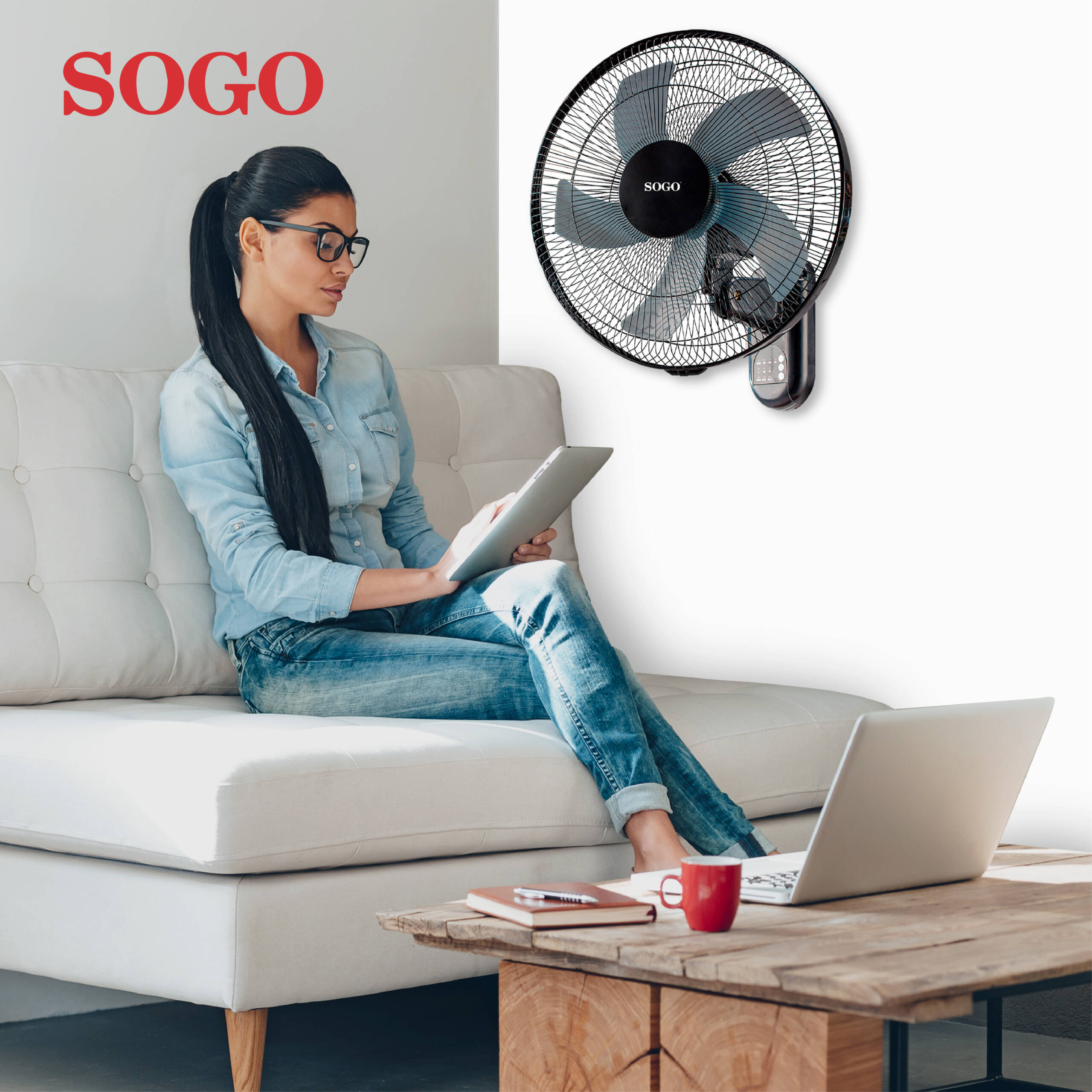 Una donna seduta su un divano usa un tablet, con un ventilatore a muro sullo sfondo.