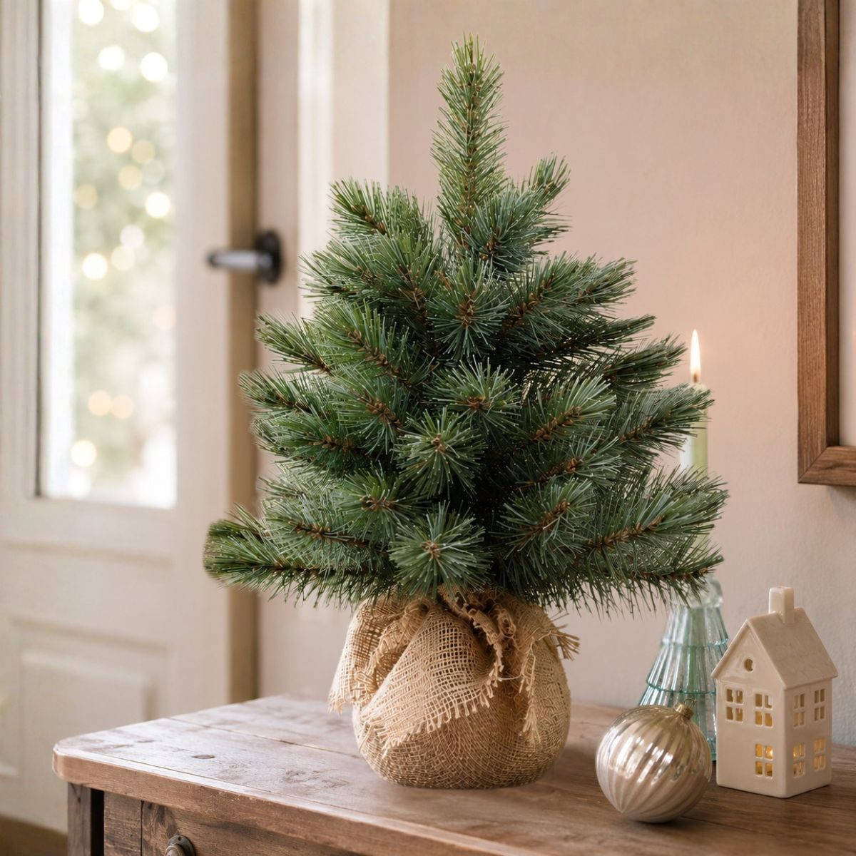 A small green pine tree in burlap sits on a wooden table with a white house light.