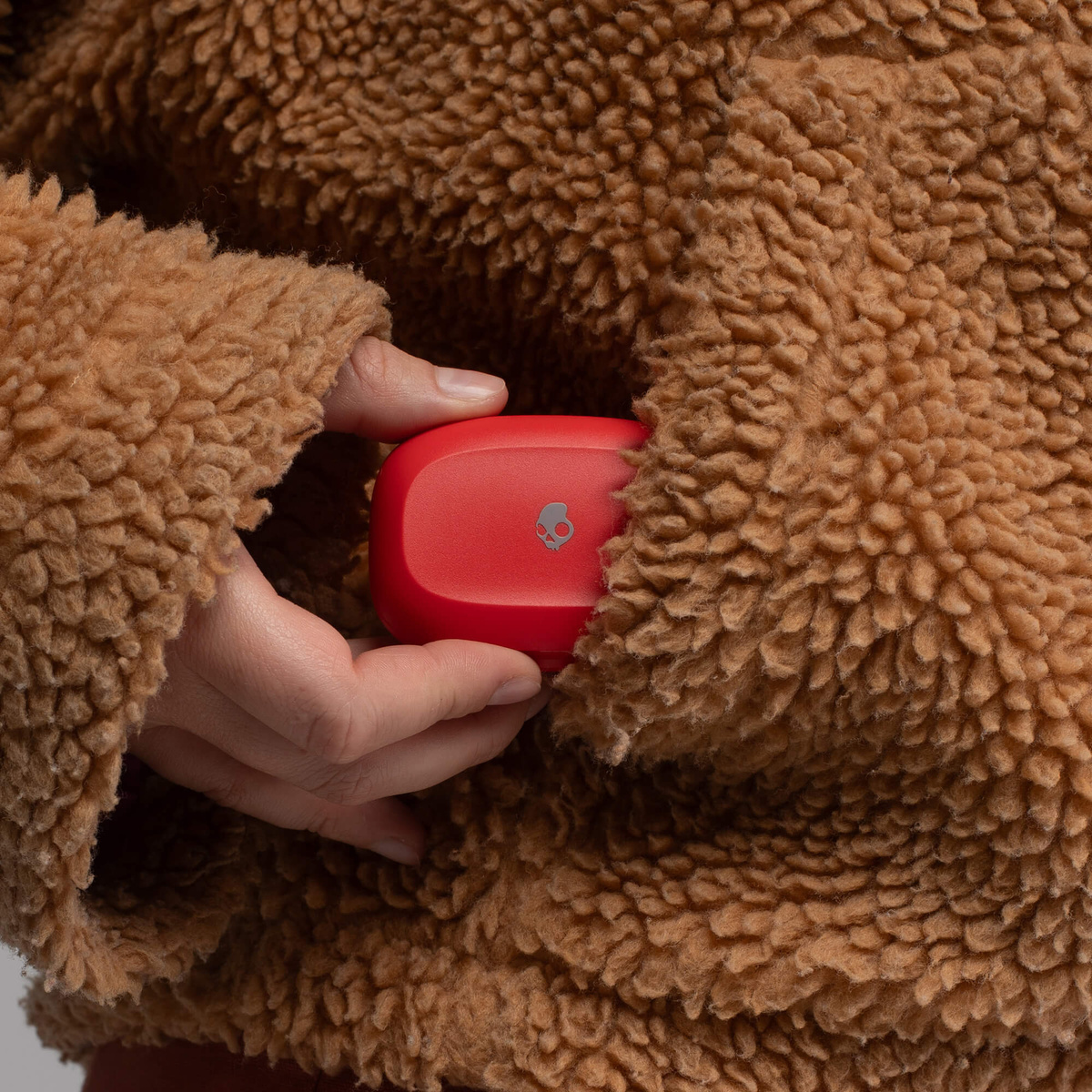 Eine Hand hält eine rote Ladehülle für Kopfhörer in einer braunen Teddyjacke.