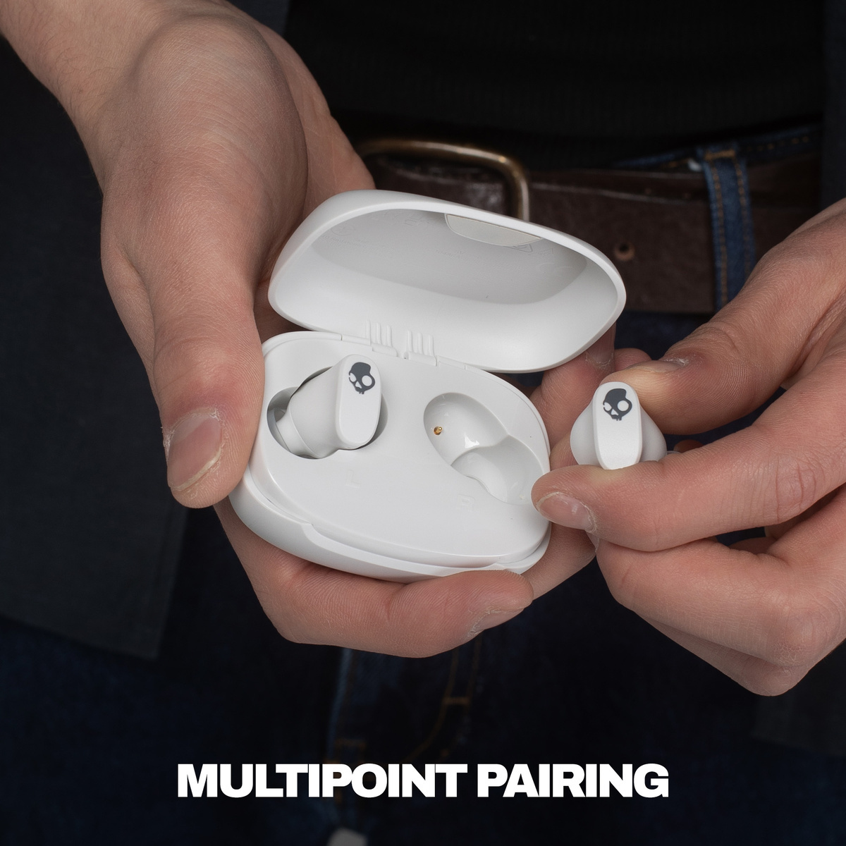 Hände halten eine weiße Skullcandy-Ohrhörerhülle mit Ohrhörern darin und einem Ohrhörer in der Hand.