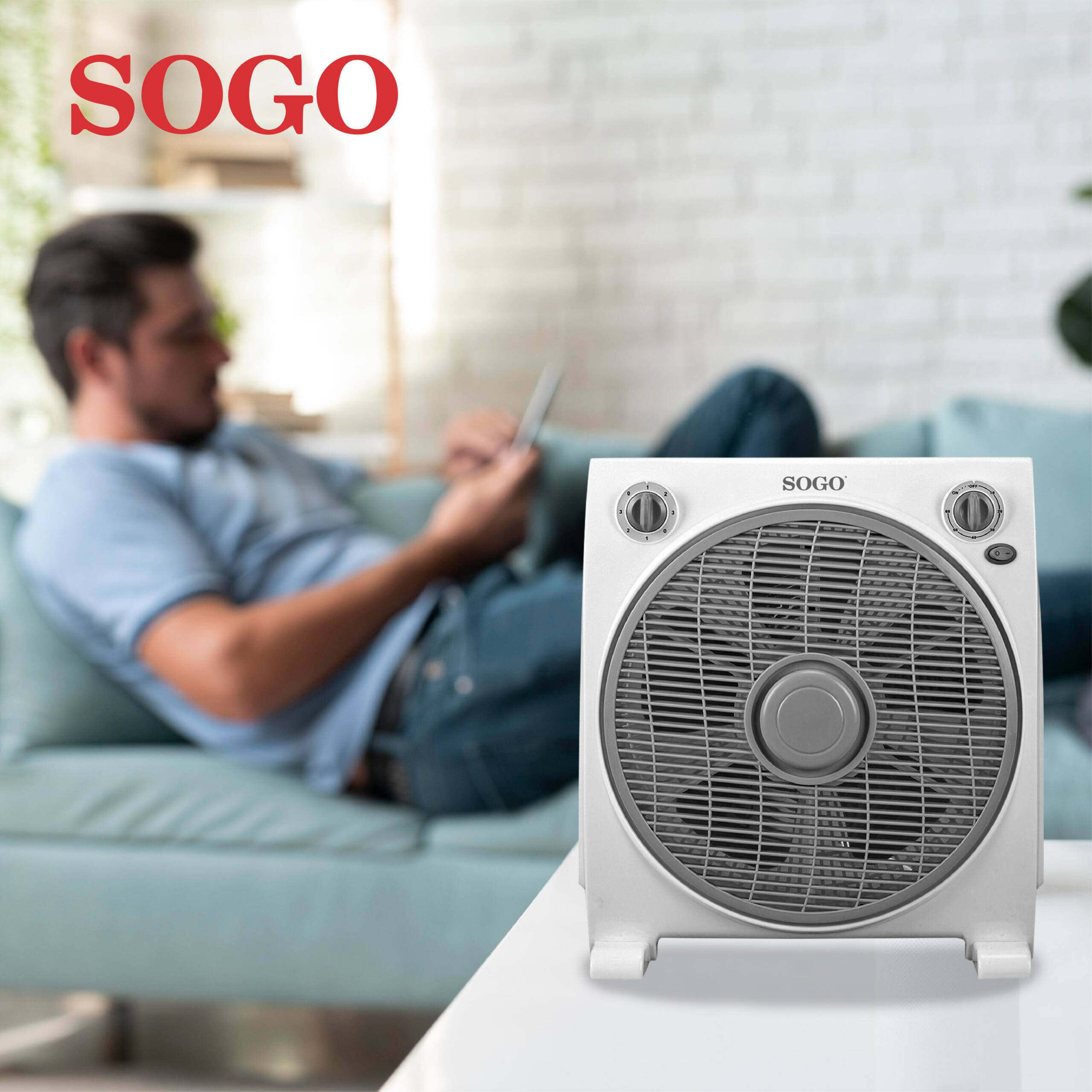 Un uomo si rilassa su un divano con un ventilatore bianco in primo piano.