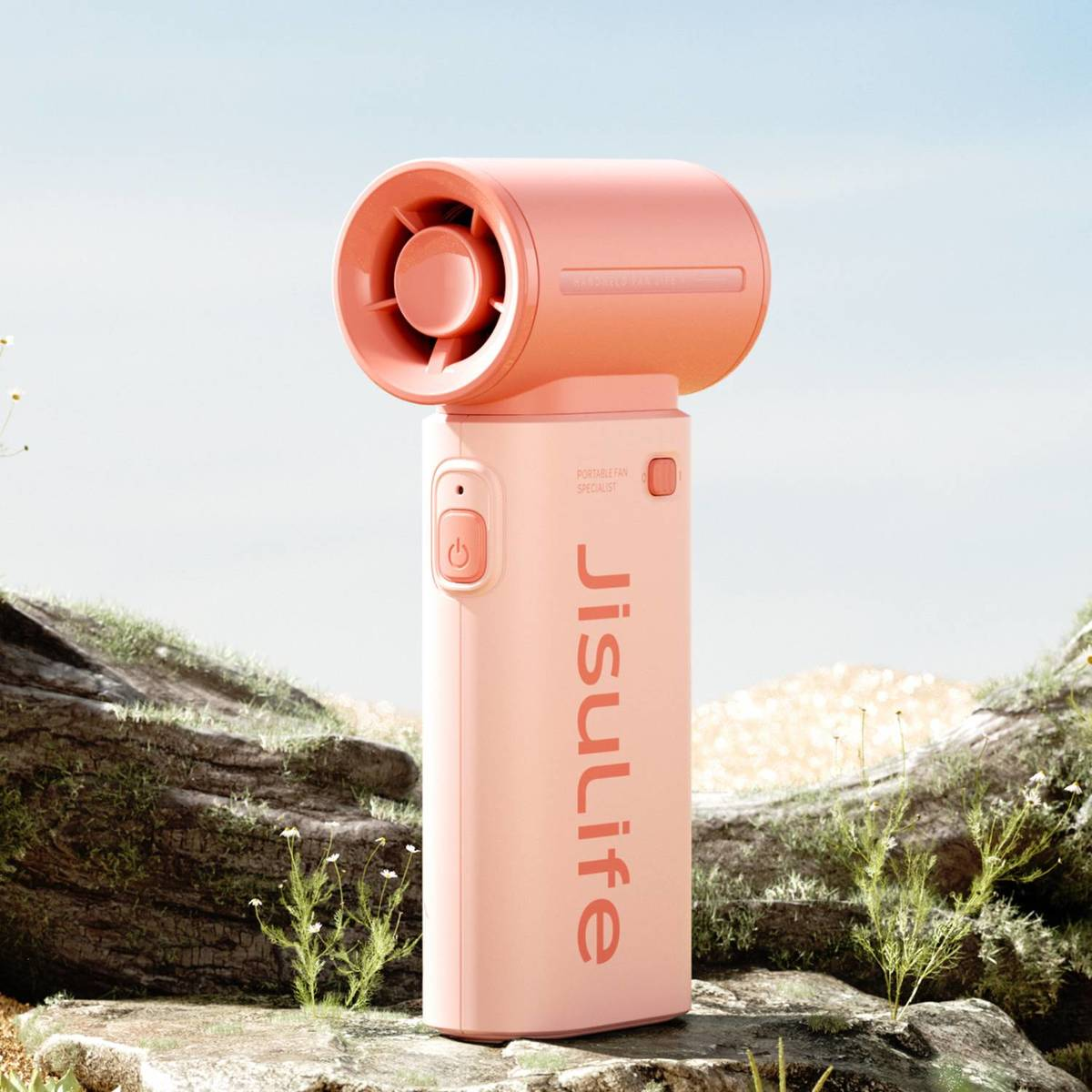 Un ventilatore portatile rosa si trova su terreno roccioso con piante e cielo azzurro.