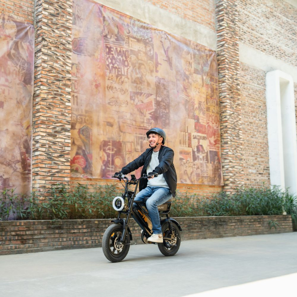 Uomo con casco guida e-bike davanti a muro di mattoni con graffiti.