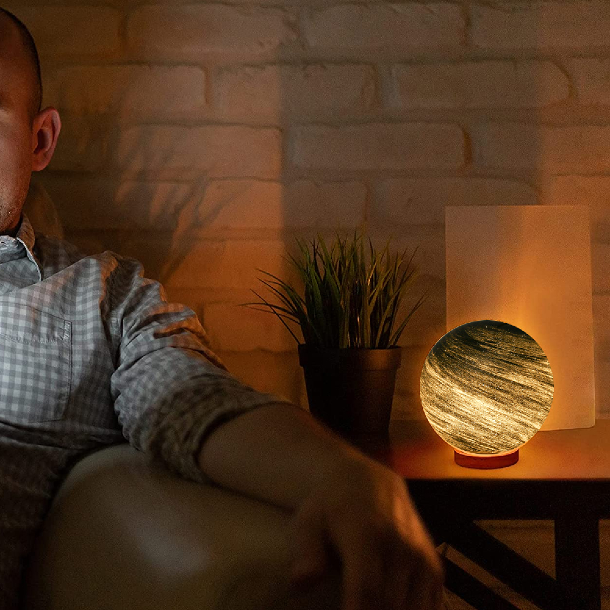 Mann sitzt auf Couch neben Tischlampe mit Muster und Topfpflanze.