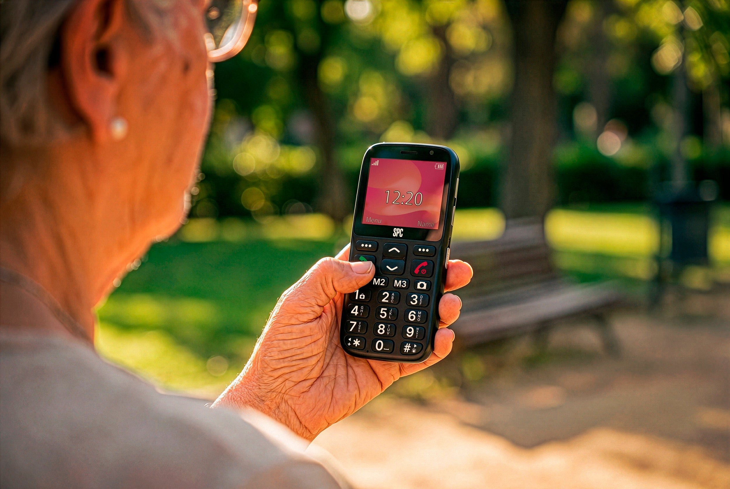 Una persona tiene un cellulare con tasti grandi all'aperto. L'ora 12:20 è visualizzata sullo schermo.