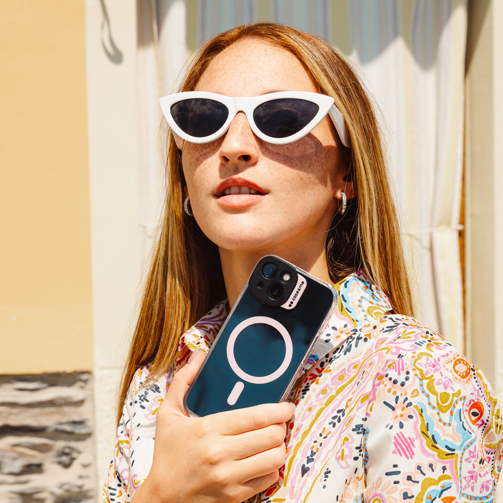 Mujer con gafas de sol sostiene teléfono con funda MagSafe ante camisa colorida.