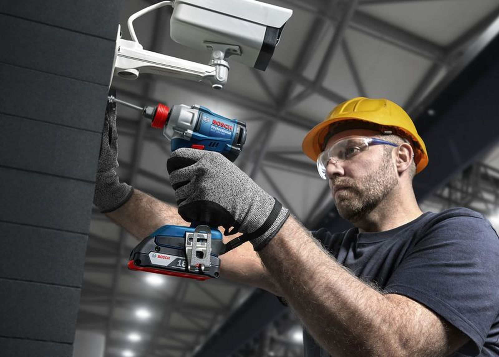 Mann in gelbem Helm montiert Kamera mit Akkuschrauber an dunkler Wand.