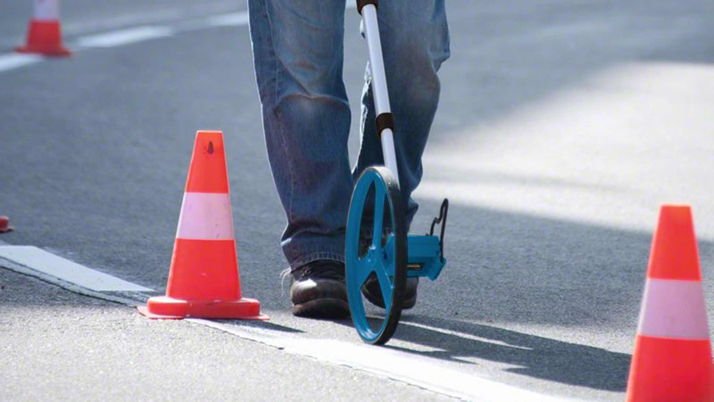 Eine Person misst mit einem Rollrad die Straße, Verkehrskegel sind aufgestellt.
