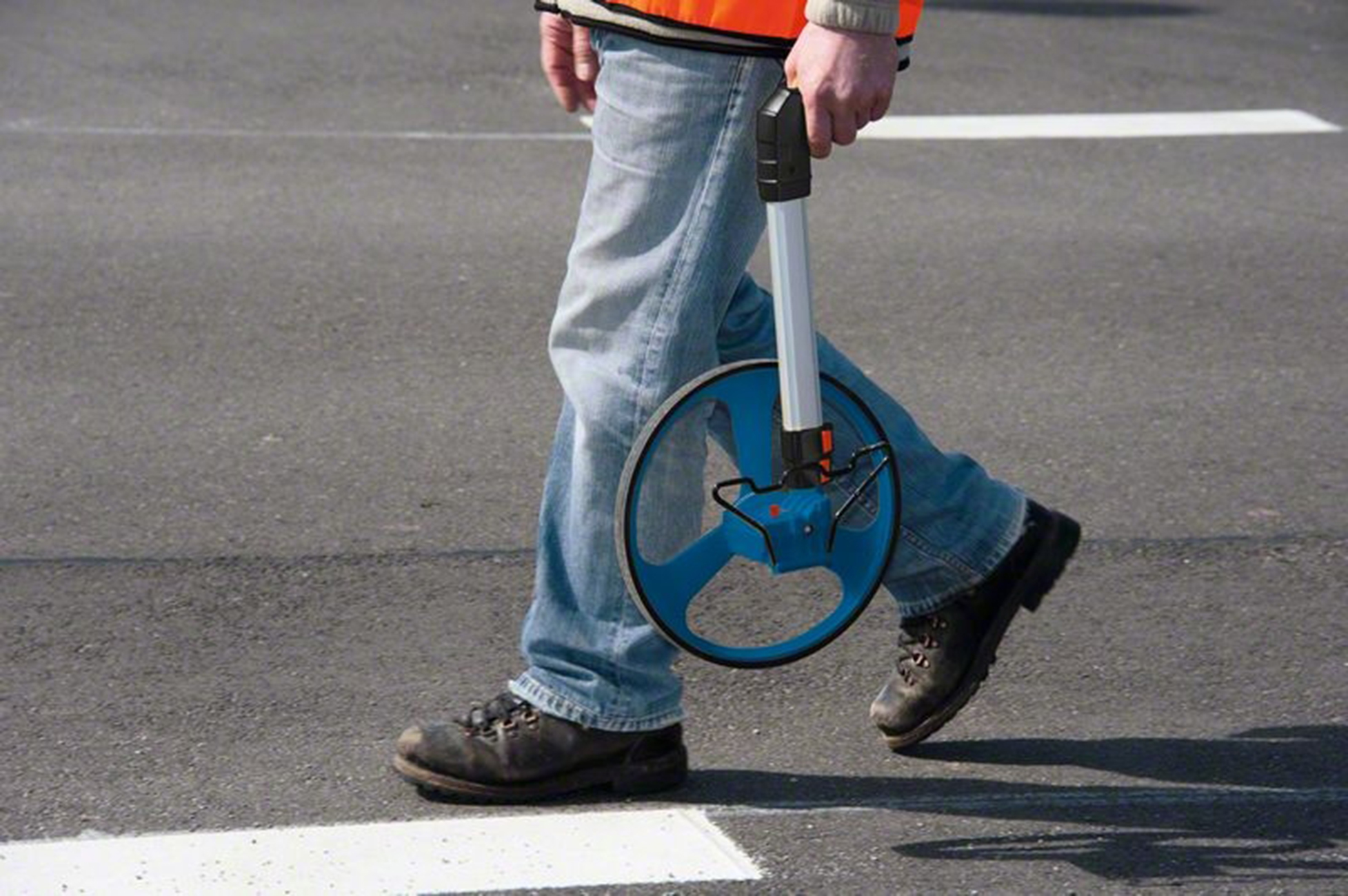 Eine Person in Jeans und orangefarbener Weste geht mit einem Messrad auf Asphalt.