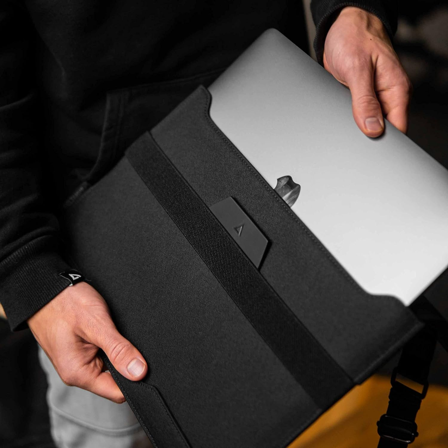 Osoba trzyma srebrnego laptopa w czarnym etui. Skupienie na dłoniach i etui.