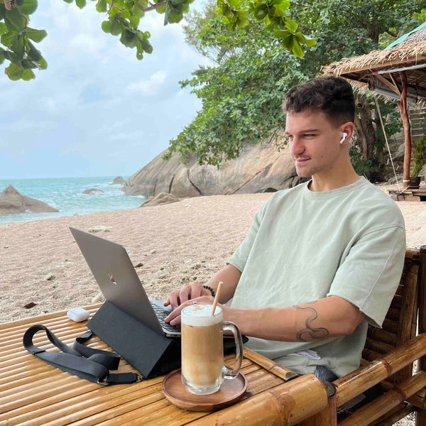 Mężczyzna pracuje na laptopie na plaży z kawą. Morze i skały w tle.