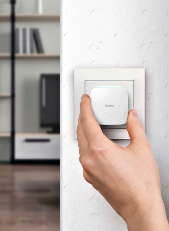 Una mano inserisce un dispositivo Hama bianco in una presa a muro bianca.