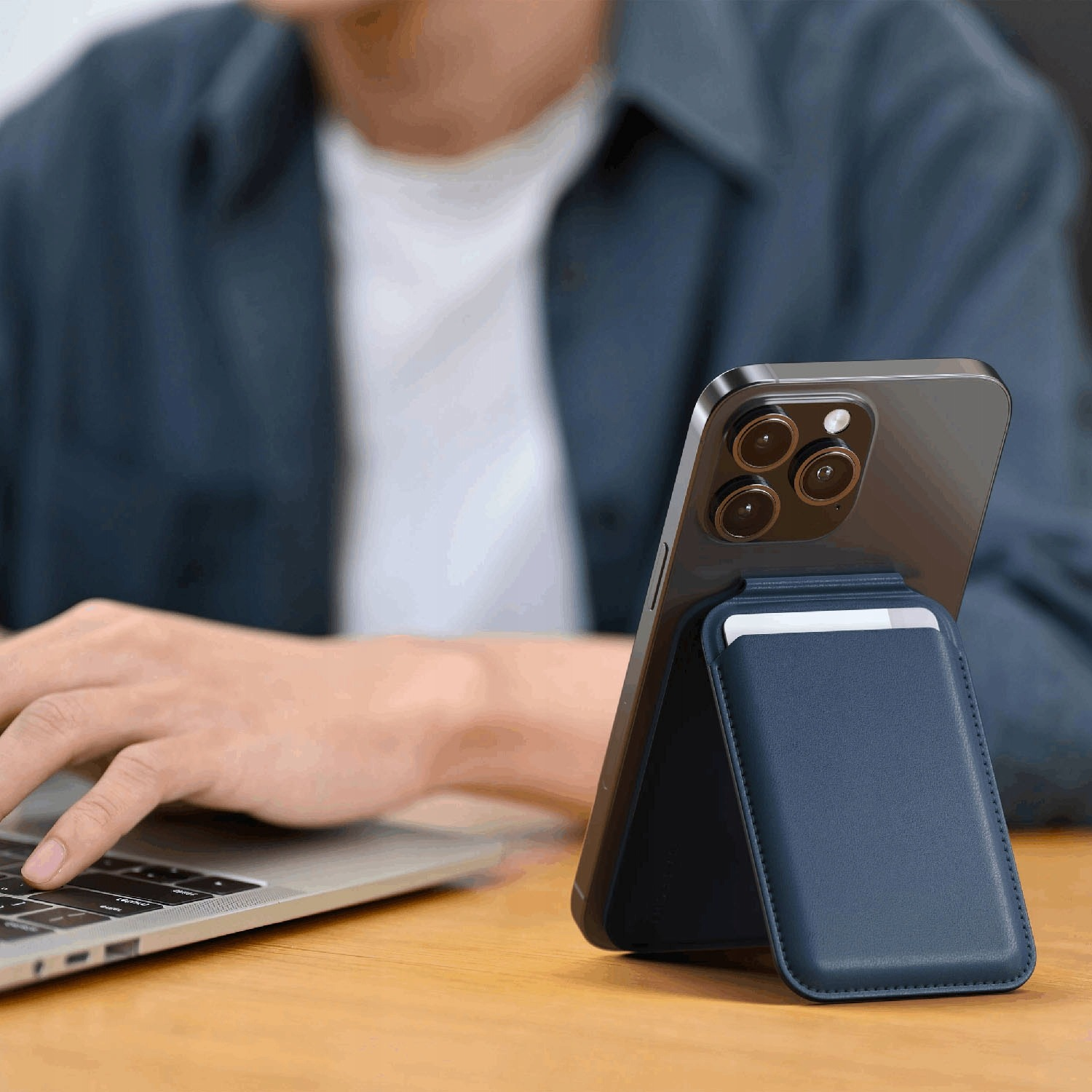 Persona digita su laptop; iPhone con porta carte blu scuro sul tavolo.