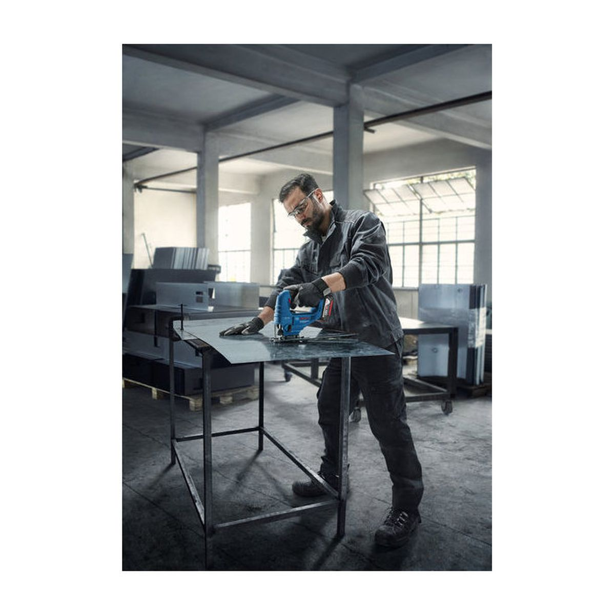 Mann in Werkstatt schneidet mit Stichsäge Metallblech auf Tisch.