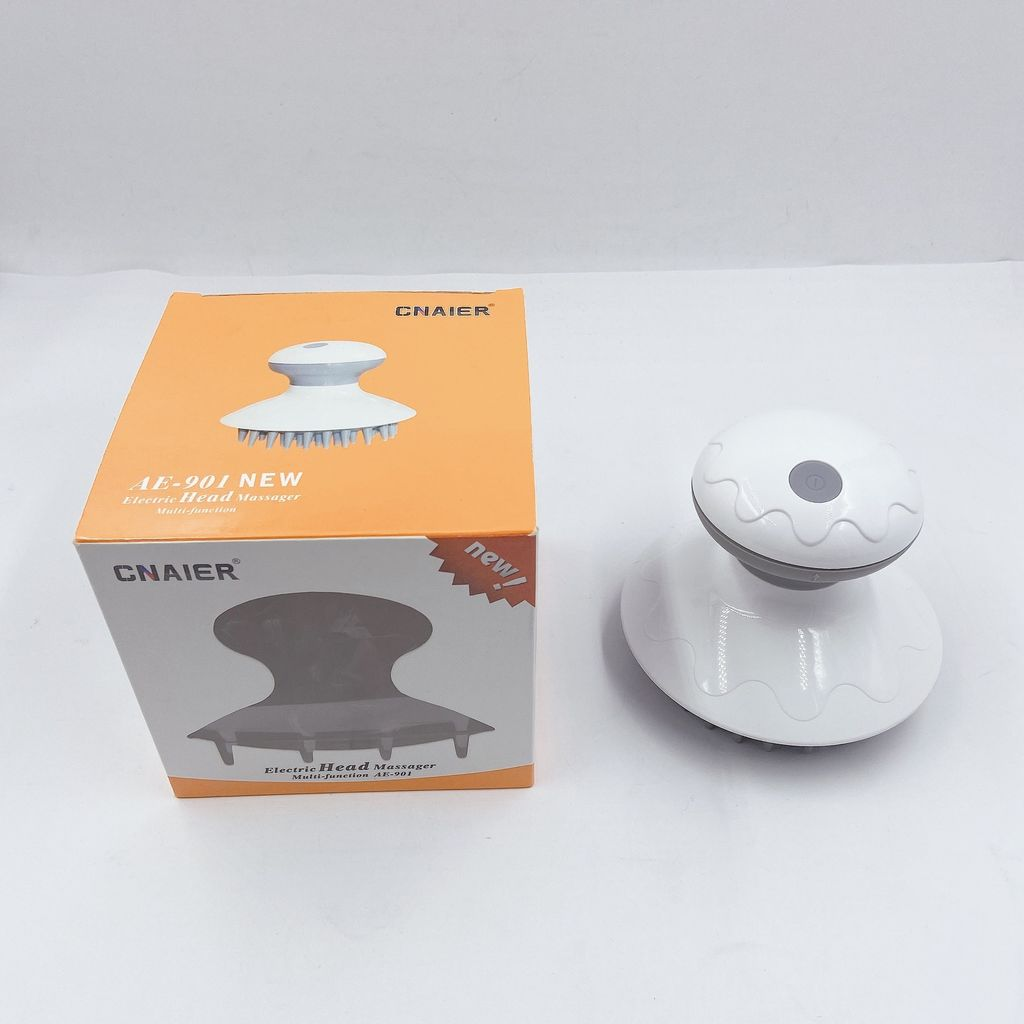Ein weißer elektrischer Kopfmassagegerät und seine orangefarbene Schachtel stehen auf einer weißen Oberfläche. Die Schachtel zeigt ein Produktbild.