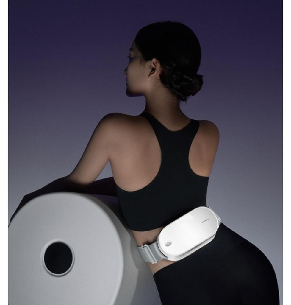 Frau in schwarzem Sport-BH und Leggings trägt ein weißes Massagegerät um die Taille.