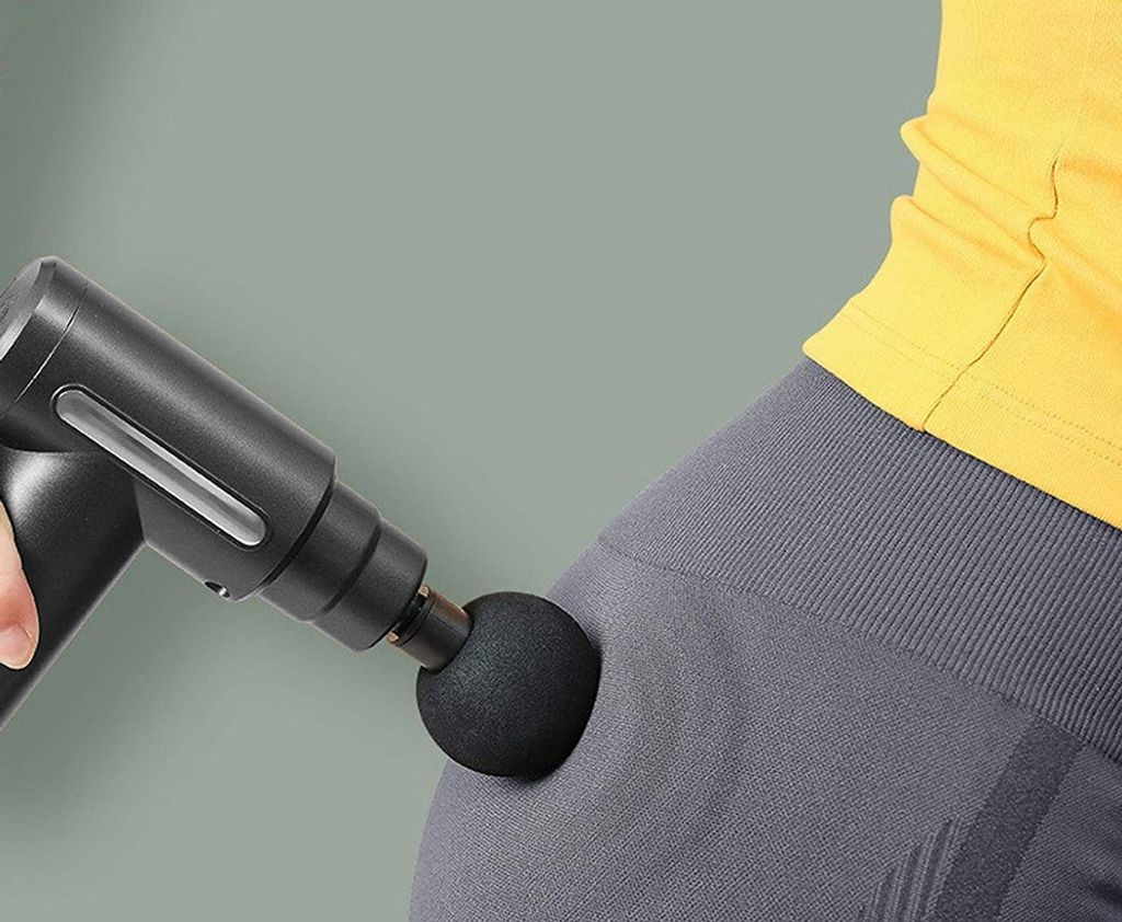 Eine schwarze Massagepistole wird auf den grau bekleideten Oberschenkel einer Person angewendet. Ein gelbes Hemd ist sichtbar.
