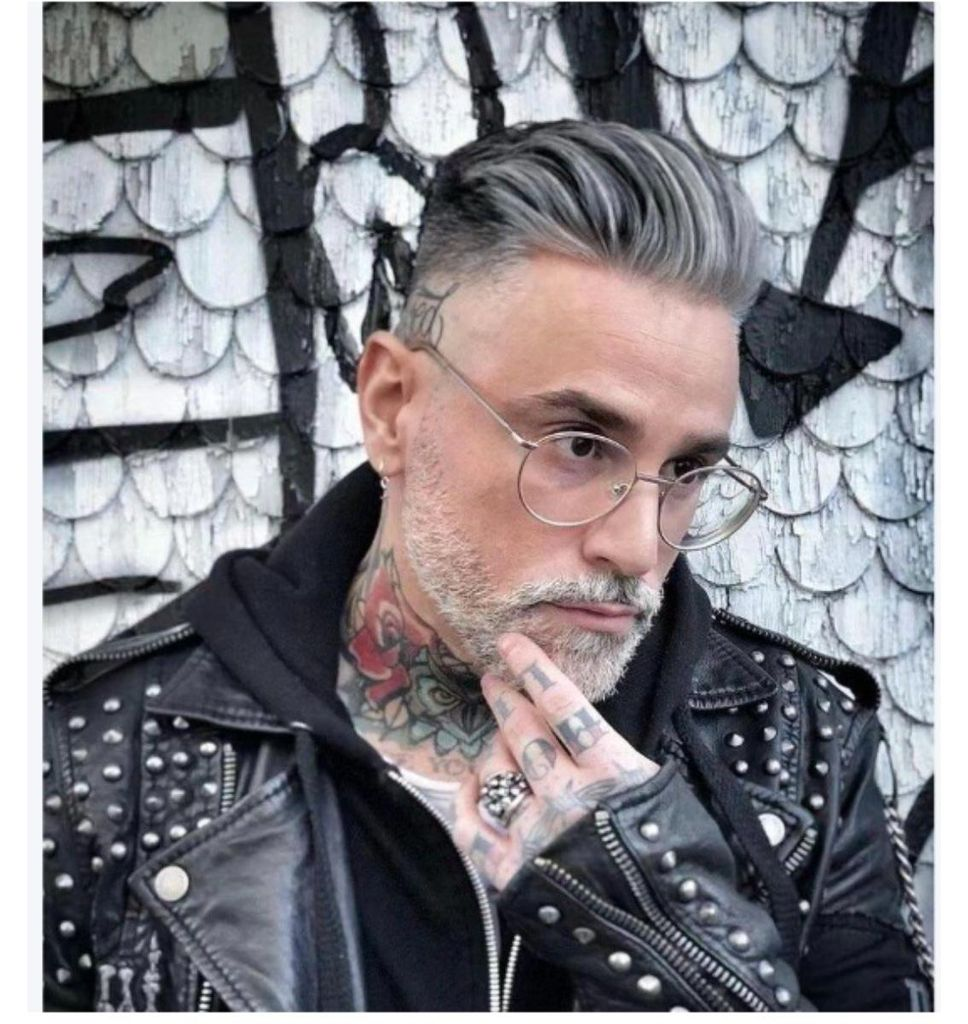 Mann mit grauem Haar, Brille, Tätowierungen und Lederjacke vor gemalter Wand.