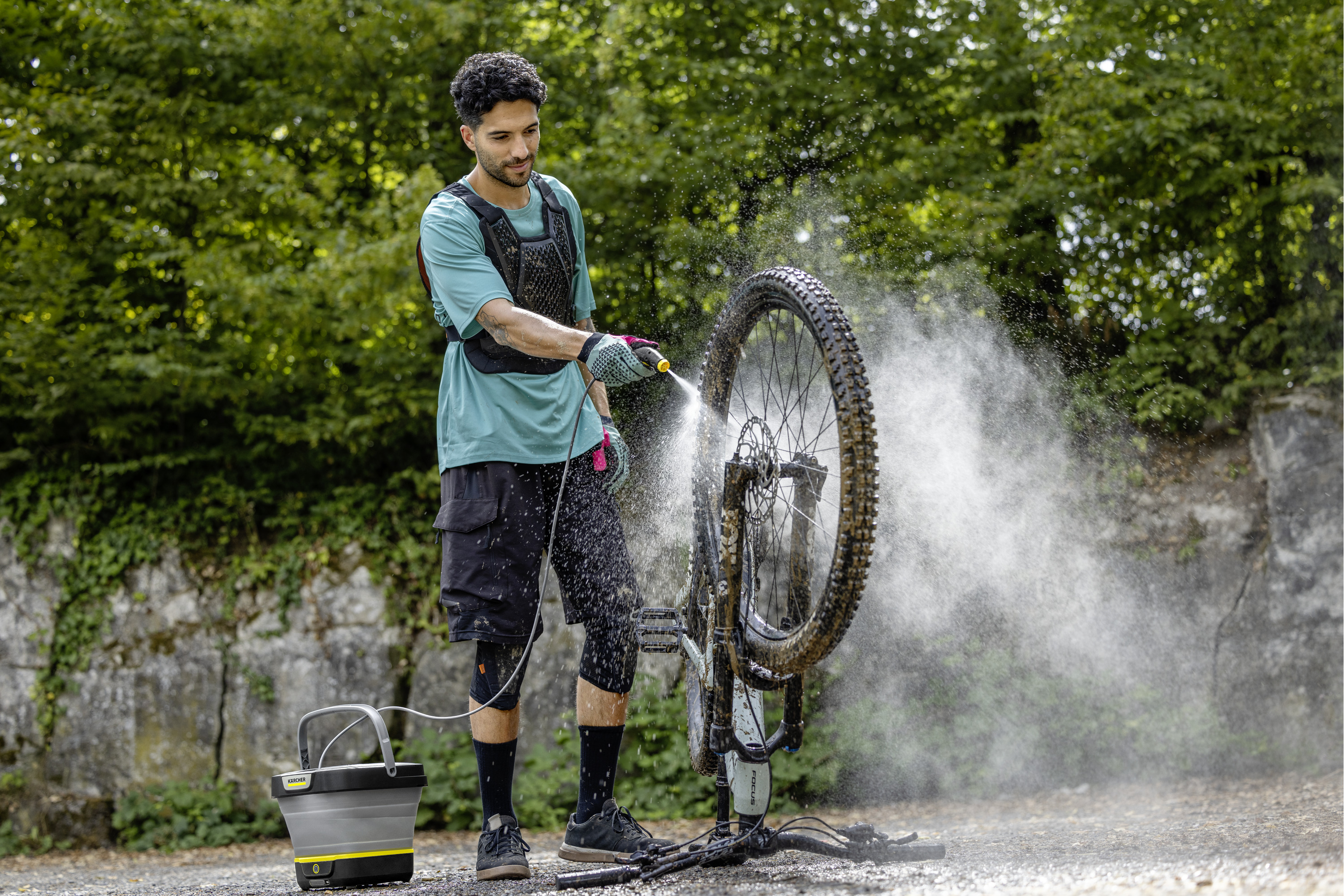 Mann wäscht Mountainbike mit Hochdruckreiniger vor Steilhang mit Bäumen im Hintergrund.