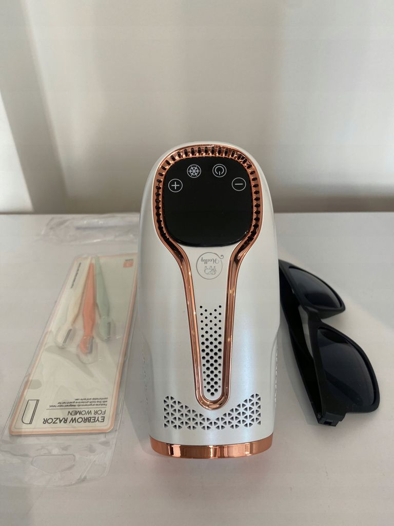 Ein weißer Haarentferner mit roségoldenen Akzenten, daneben eine Augenbrauenrasierverpackung und Sonnenbrille.