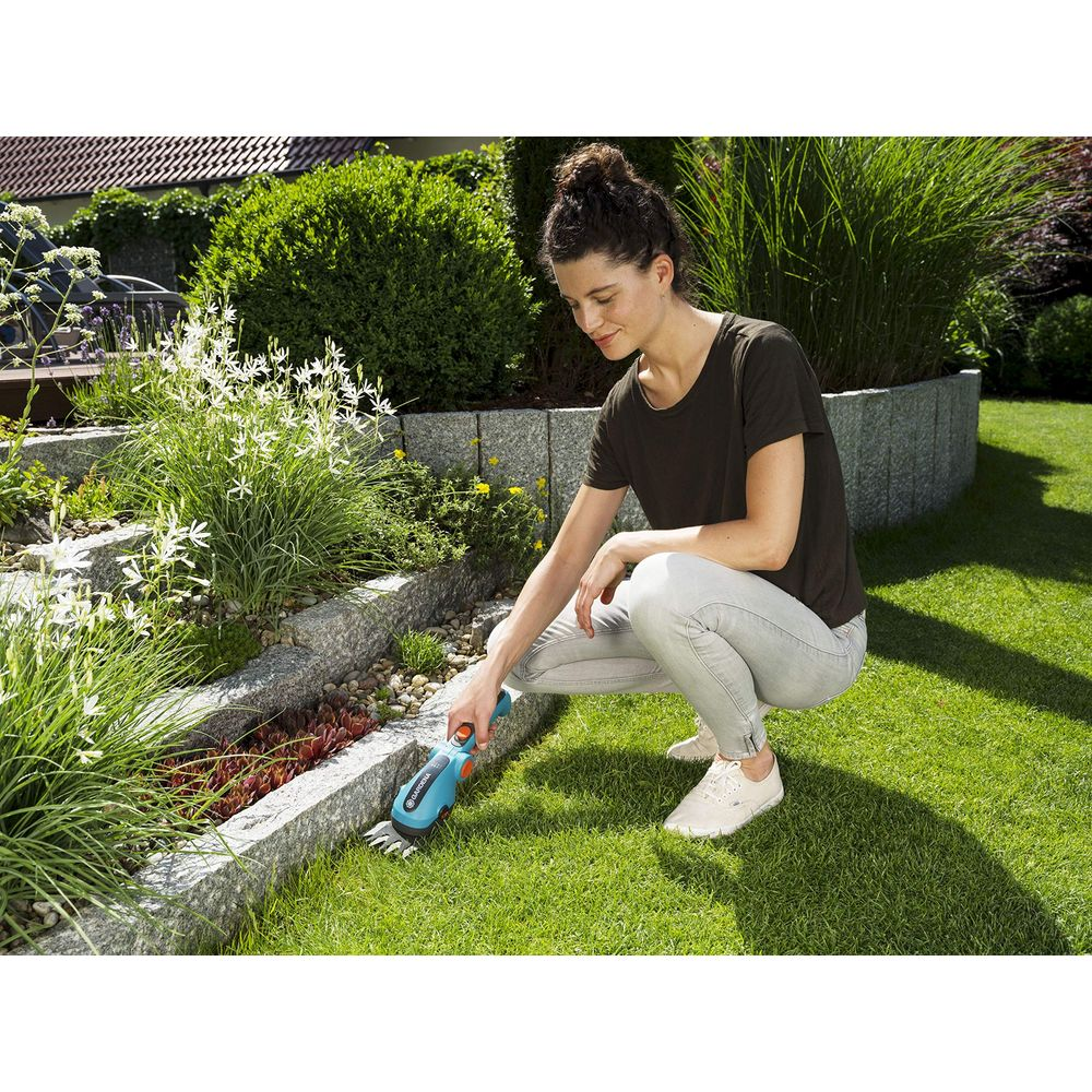 Eine Frau kniet im Garten und trimmt mit einem kleinen Gerät den Rasen.