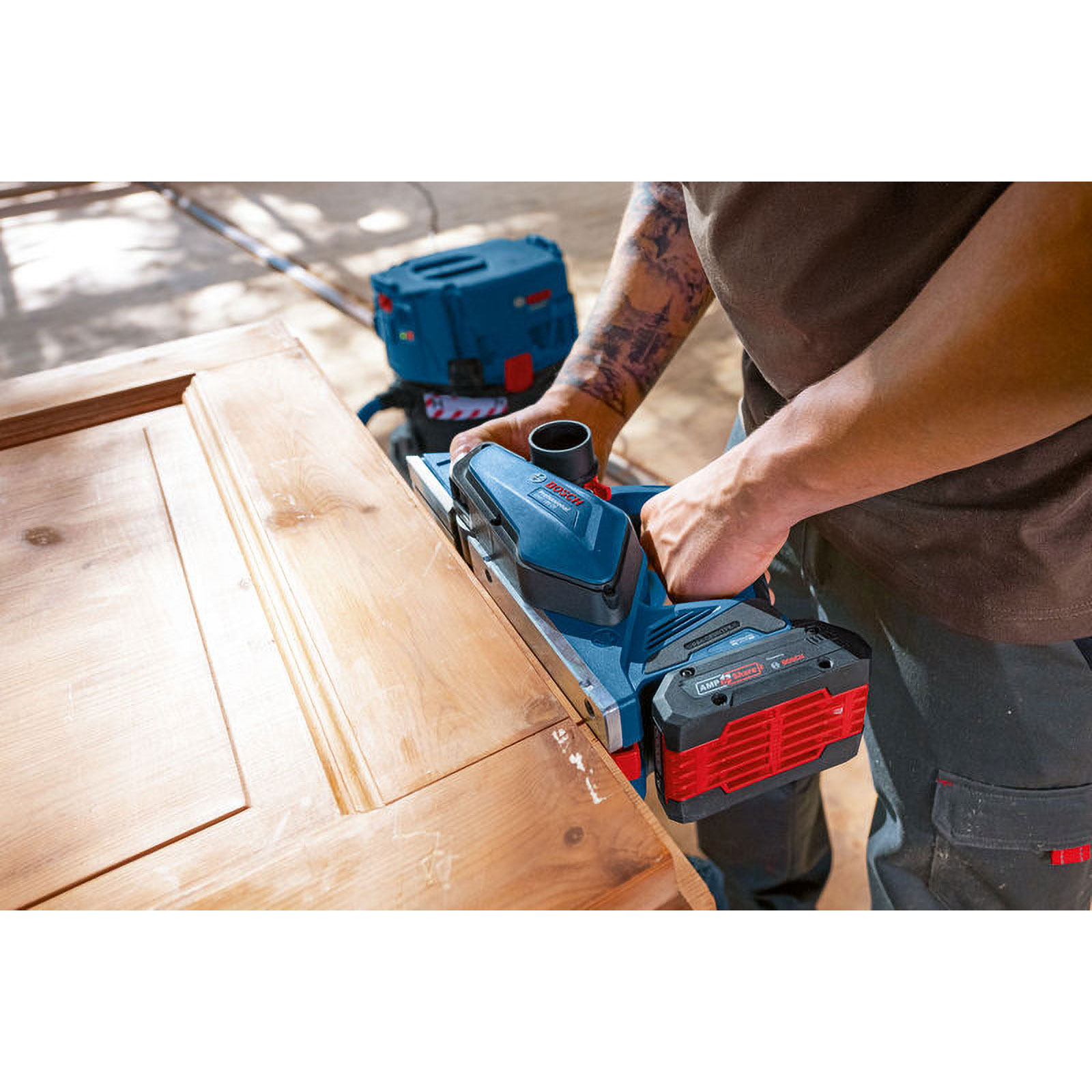 Ein Mann bearbeitet eine Holztür mit einem blauen und roten Elektrohobel. Im Hintergrund ein blauer Kasten.