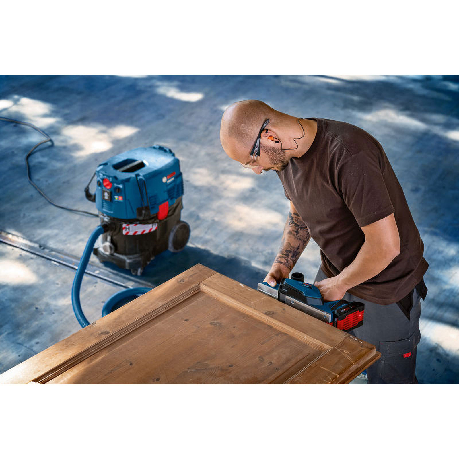 Mann mit Schutzbrille bearbeitet Holztür mit Hobel, Staubsauger daneben.