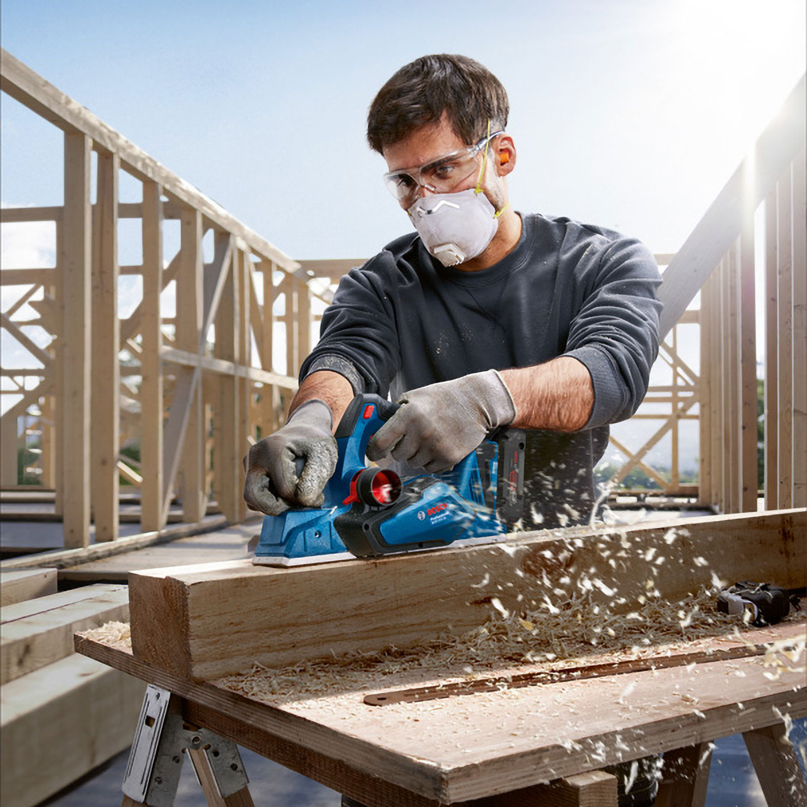 Mann hobelt Holz mit blauer Elektrowerkzeug vor Holzgerüst.
