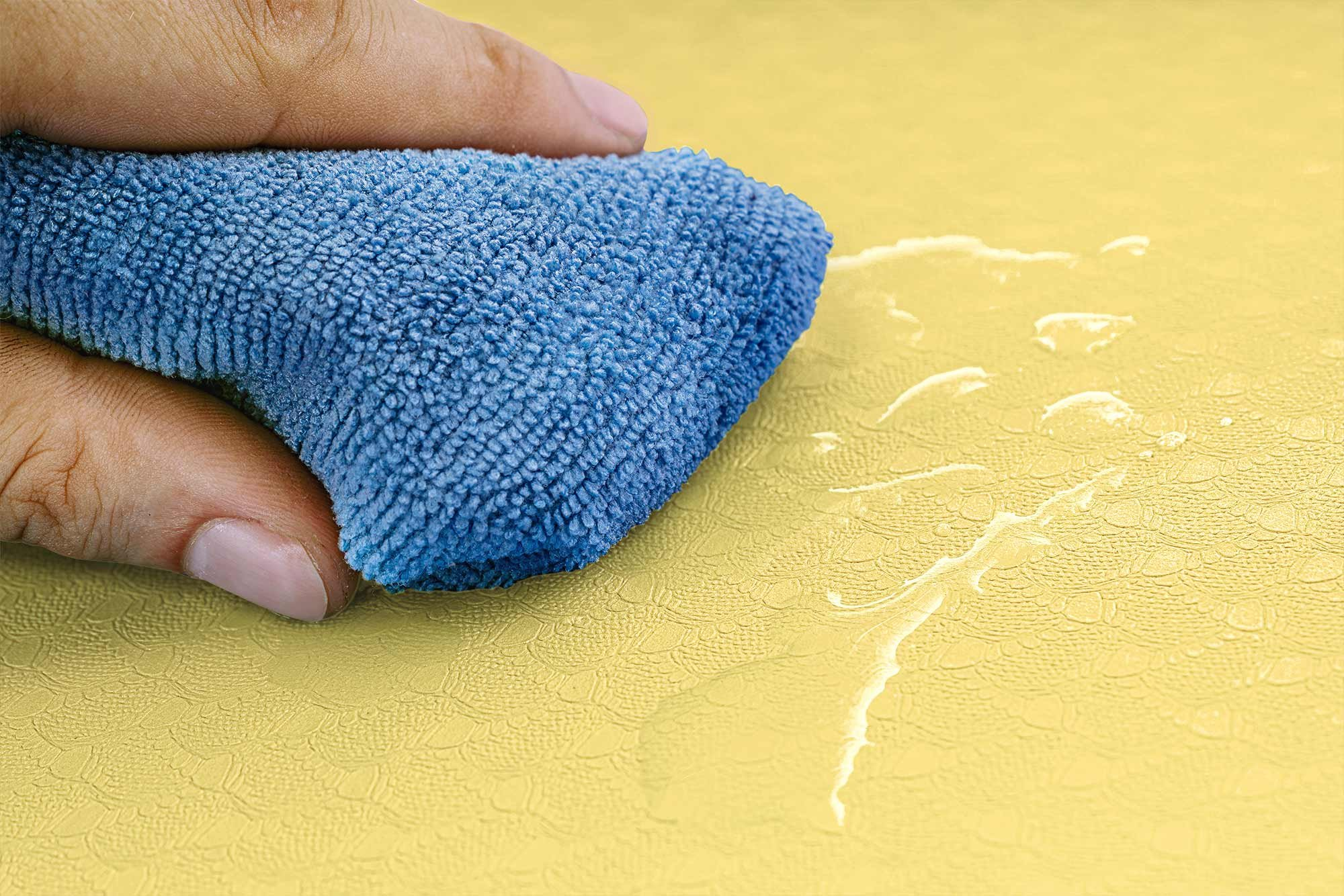 Eine Hand hält einen blauen Lappen, der über eine gelbe Oberfläche wischt.