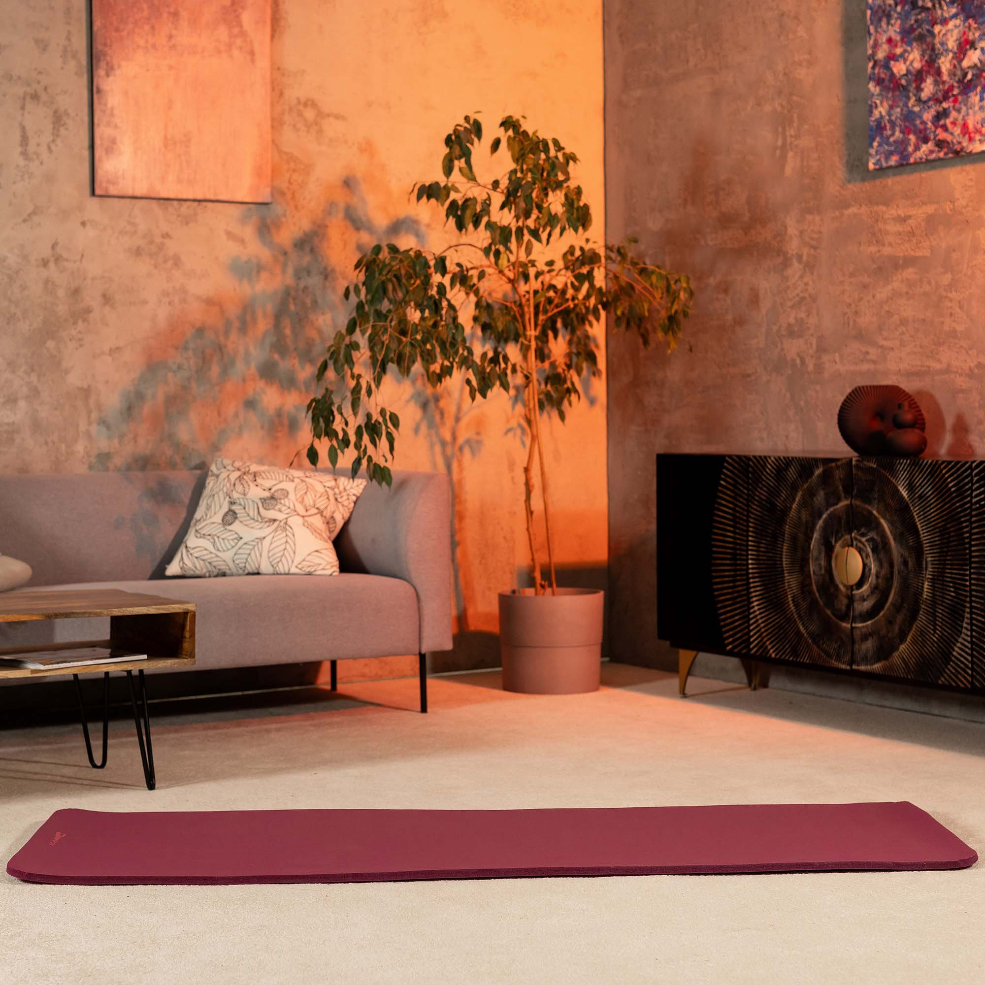 Ein dunkelrotes Yogamatte liegt auf einem hellen Teppich vor einem Sofa.