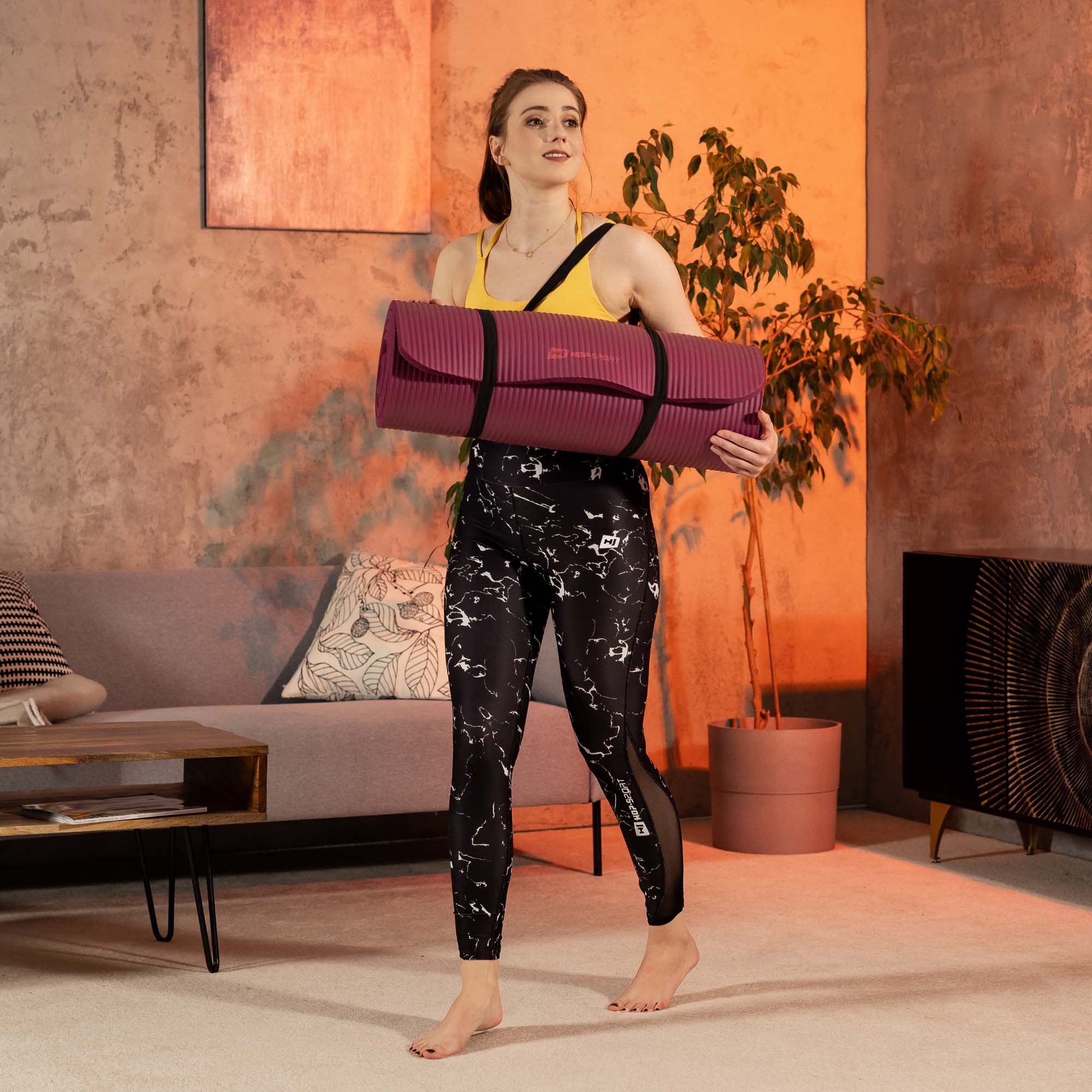 Eine Frau trägt eine lila Yogamatte, gelbes Top, gemusterte Leggings.
