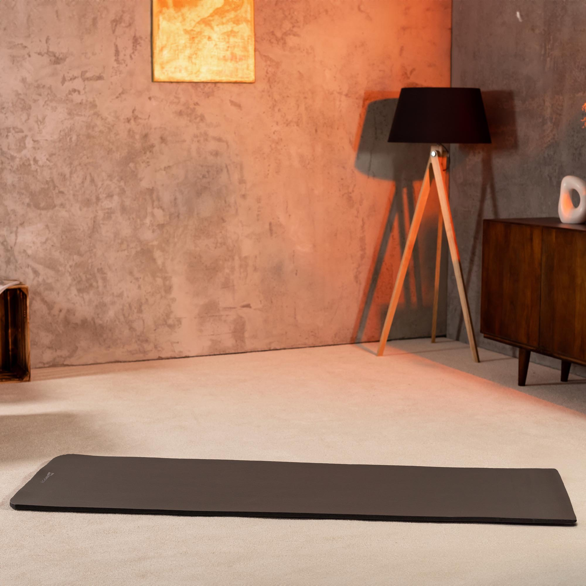 Eine dunkle Yogamatte liegt auf einem hellen Teppich vor einer Wand mit Kunst und Lampe.