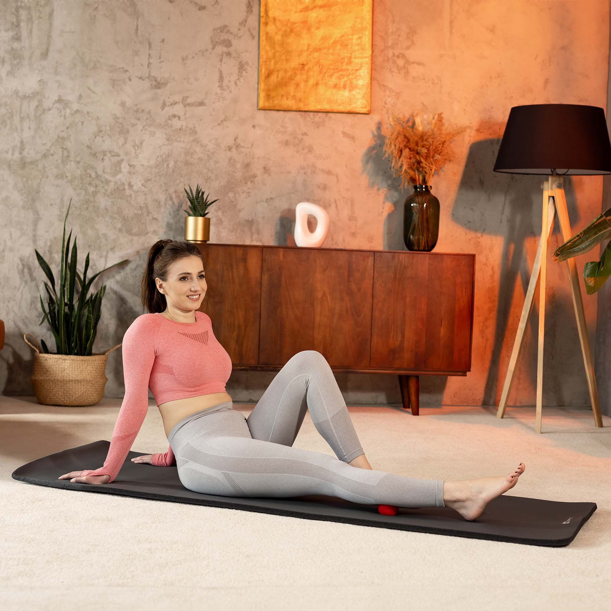 Frau in pinkem Oberteil und grauer Leggings auf Matte, massiert Bein mit rotem Ball.