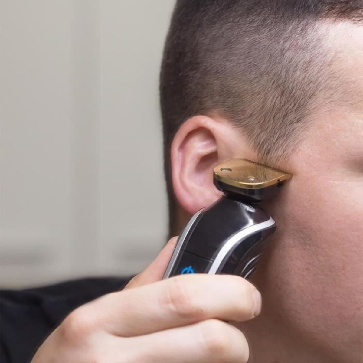Nahaufnahme: Eine Person rasiert sich mit einem schwarzen und goldenen Trimmer die Haare neben dem Ohr.
