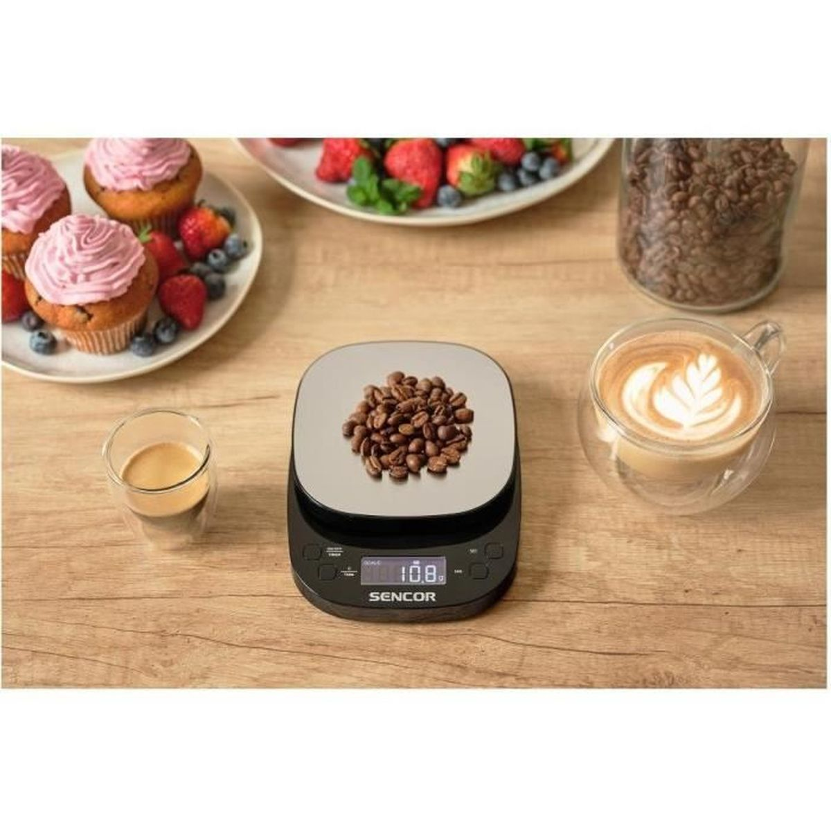 Eine digitale Waage mit Kaffeebohnen, daneben Becherkuchen, Beeren und Latte Art Kaffee.