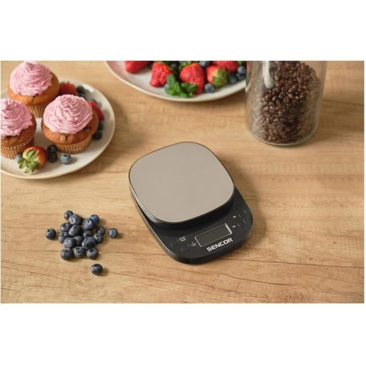 Eine digitale Küchenwaage steht auf einem Holztisch neben Beeren und Kuchen.