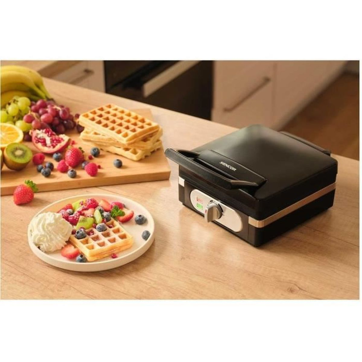 Ein schwarzer Waffelmaker neben Waffeln und Früchten auf einem Holztisch.