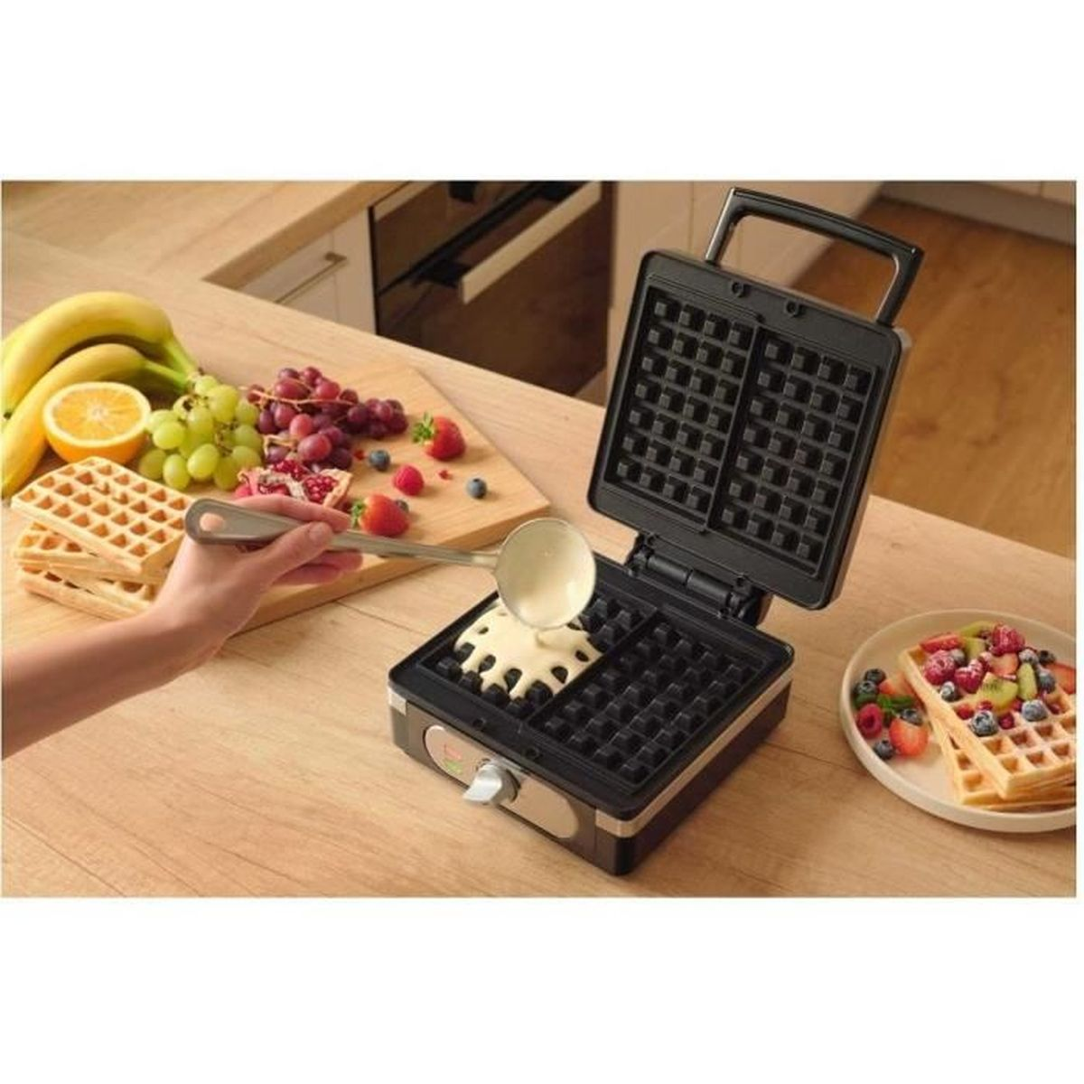 Eine Hand gießt Teig in eine offene Waffelmaschine. Früchte und Waffeln liegen daneben.