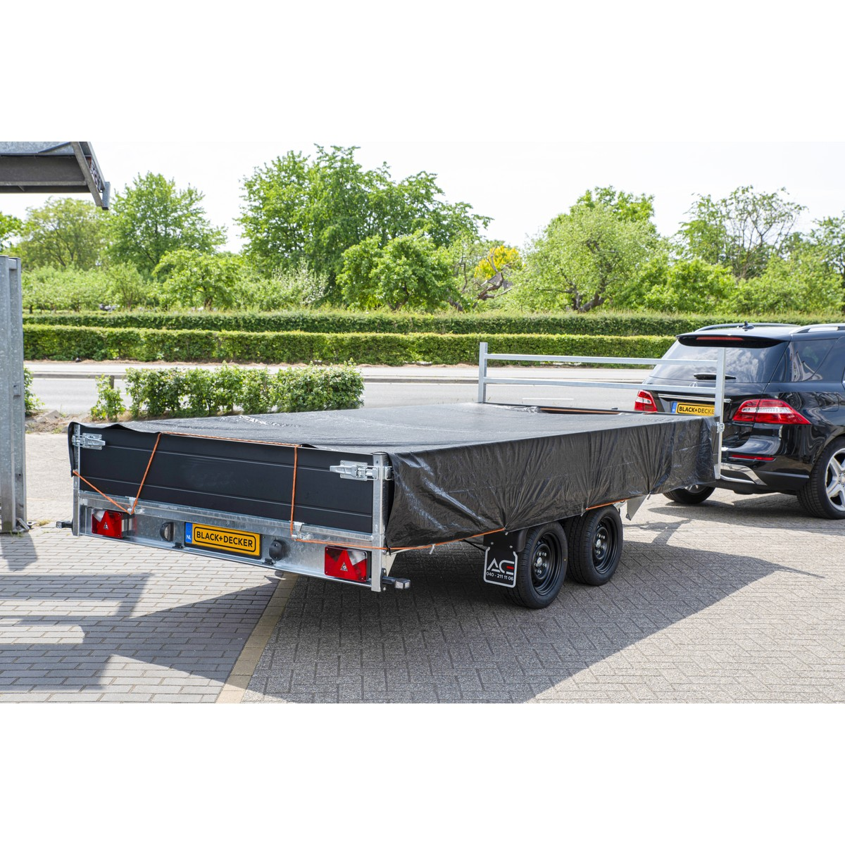 Een zwarte trailer met zwart zeil, vastgezet met touwen, is aan een zwarte auto gekoppeld.