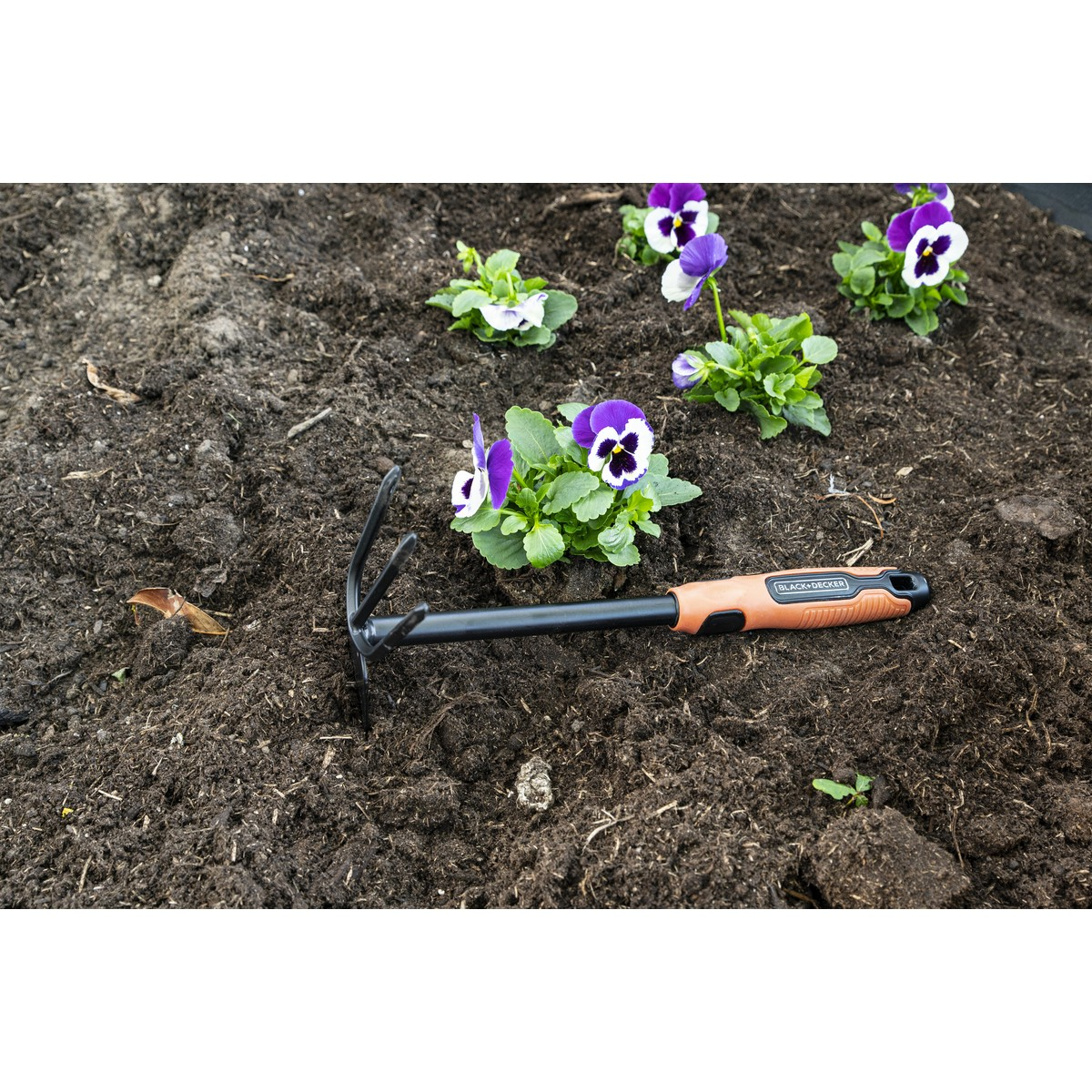 A black and orange hand rake rests on dark soil beside purple and white pansies.